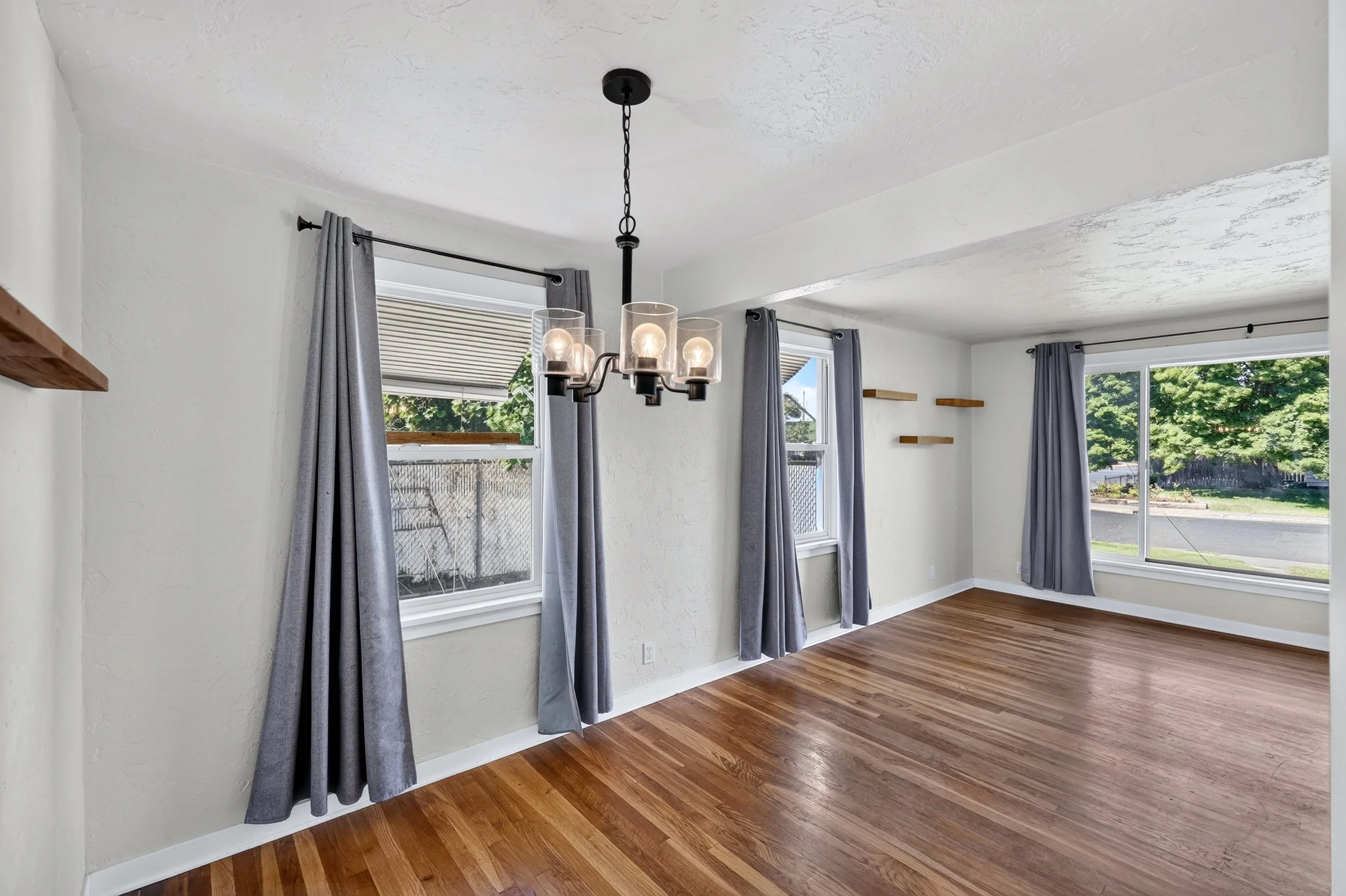 Empty room with hardwood floors, white textured walls, four windows with gray curtains, and a black chandelier with glass shades hanging from the ceiling.