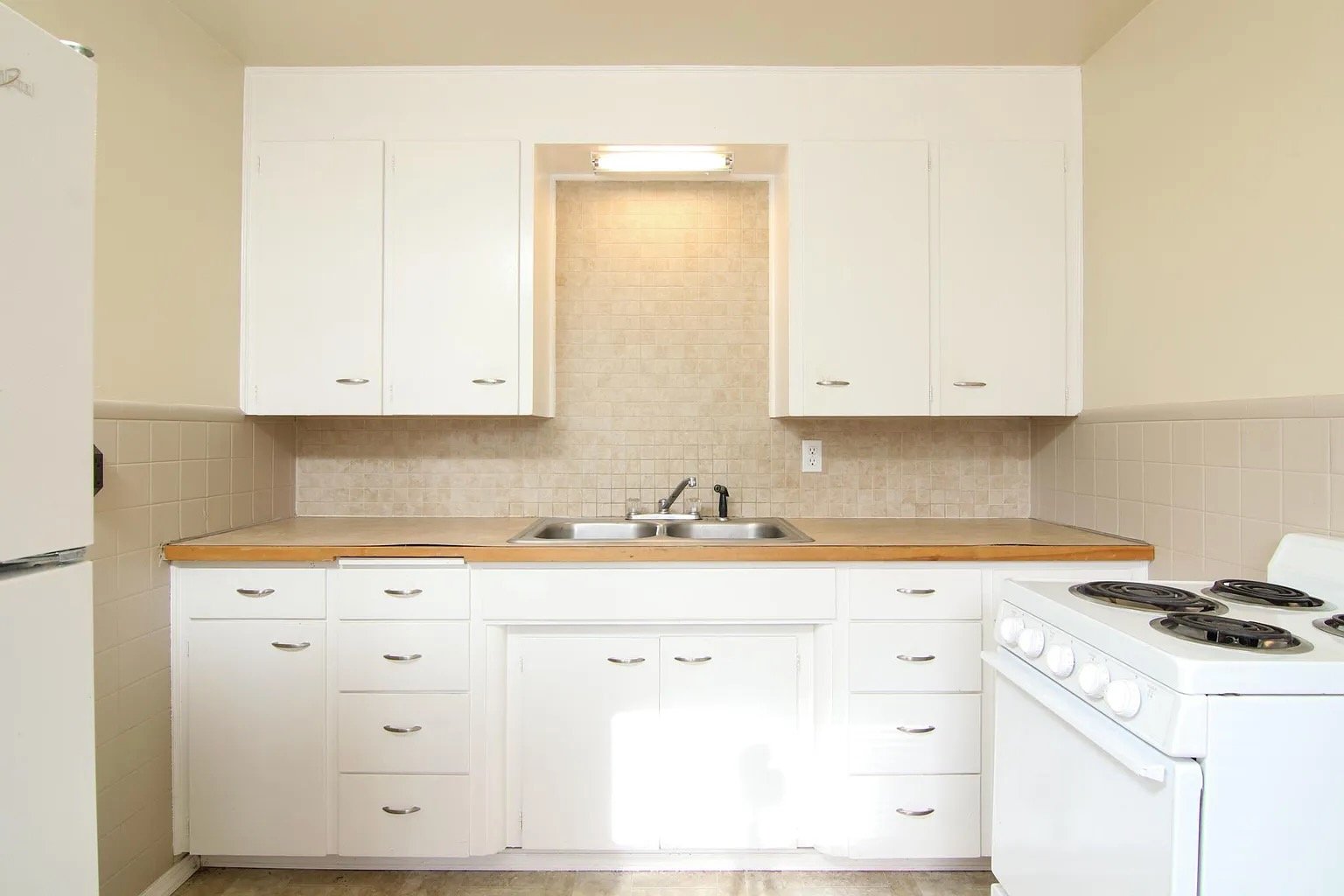 Kitchen with white cabinets, a beige tiled backsplash, a double sink, a stove, and a partial view of a refrigerator.