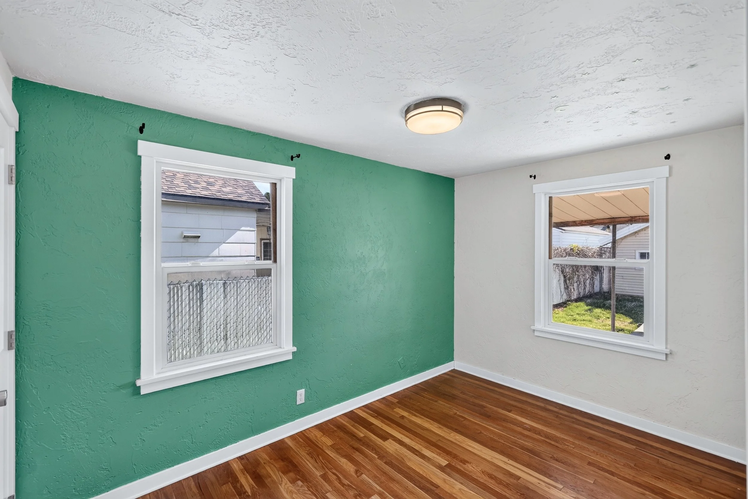 Room with green and white painted walls, two windows, wooden floor, ceiling light, and visible outdoor yard.
