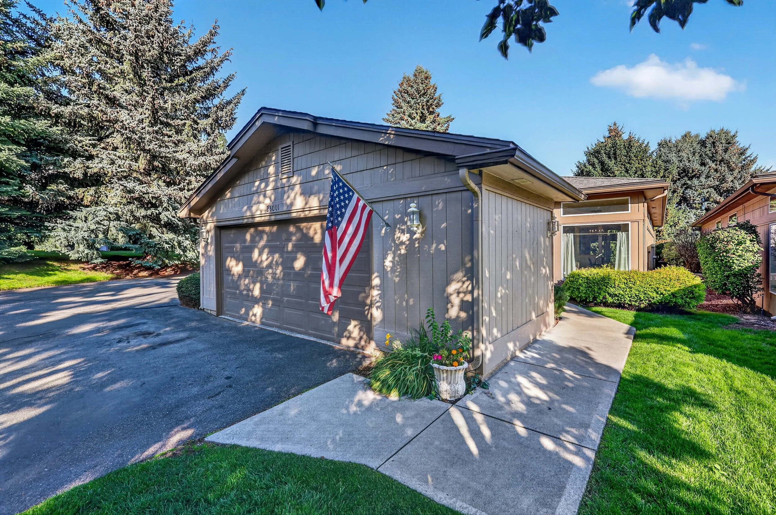 A suburban house with a attached garage, beige exterior, and an American flag on a pole in front. The yard has green grass, bushes, and a flower pot, with a clear blue sky and trees in the background.