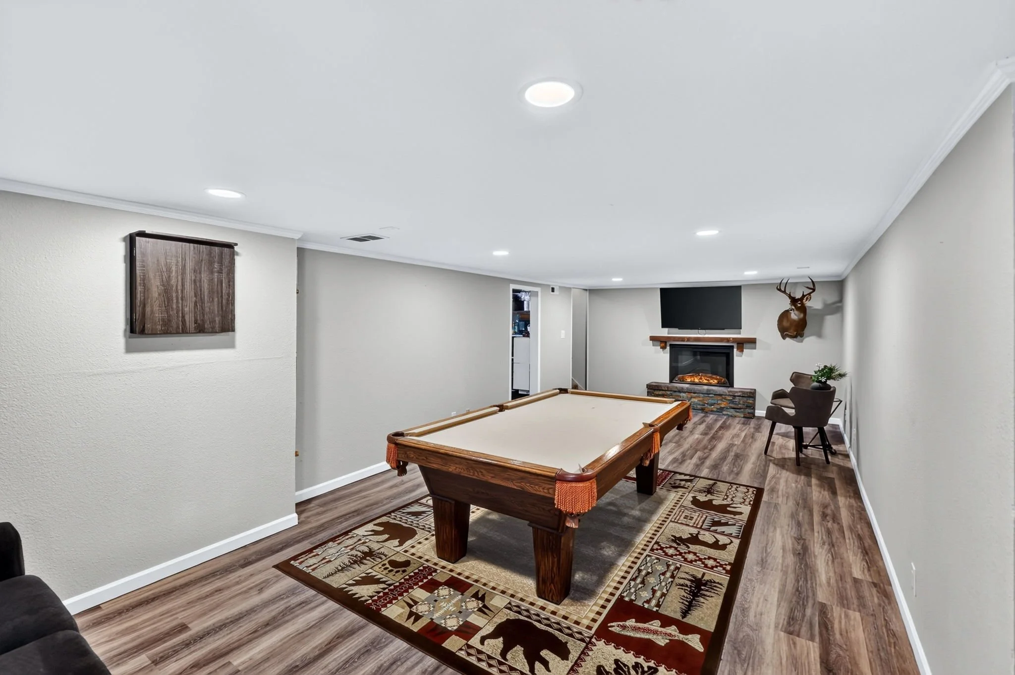 A spacious basement with a pool table, a fireplace, and a mounted deer head, with wood flooring and a patterned area rug.
