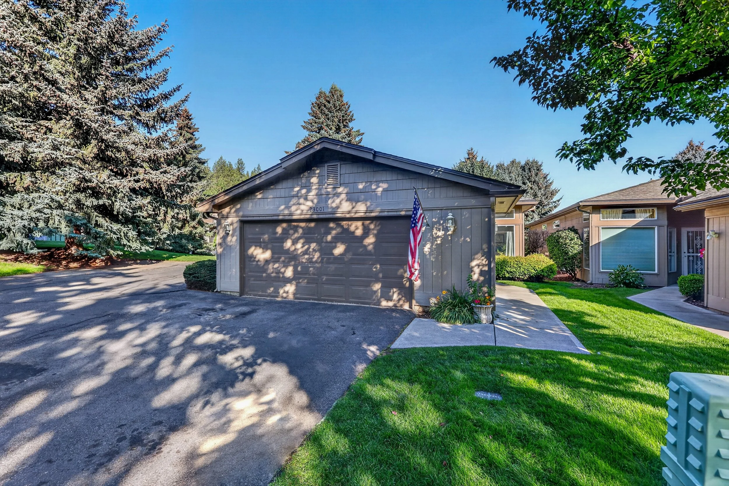 A suburban house with a gray front door, a garage, American flag, green lawn, and trees on a sunny day.