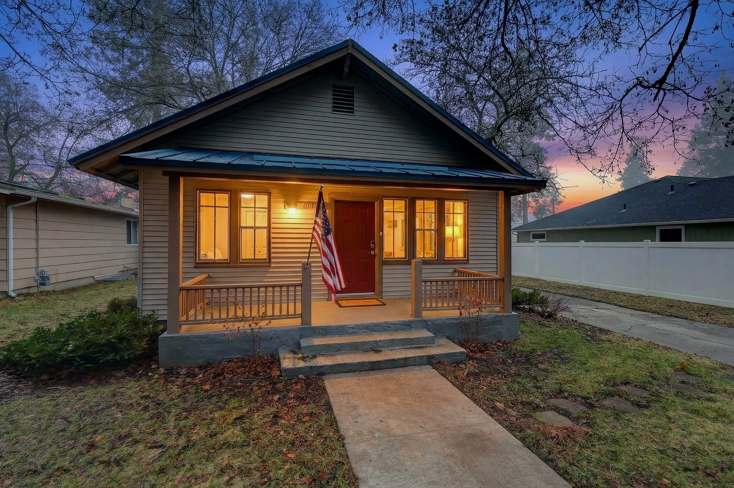 Front view of a cozy house at sunset with a well-lit interior, a small porch with steps, an American flag hanging, and a walkway leading to the sidewalk, surrounded by a lawn and trees.
