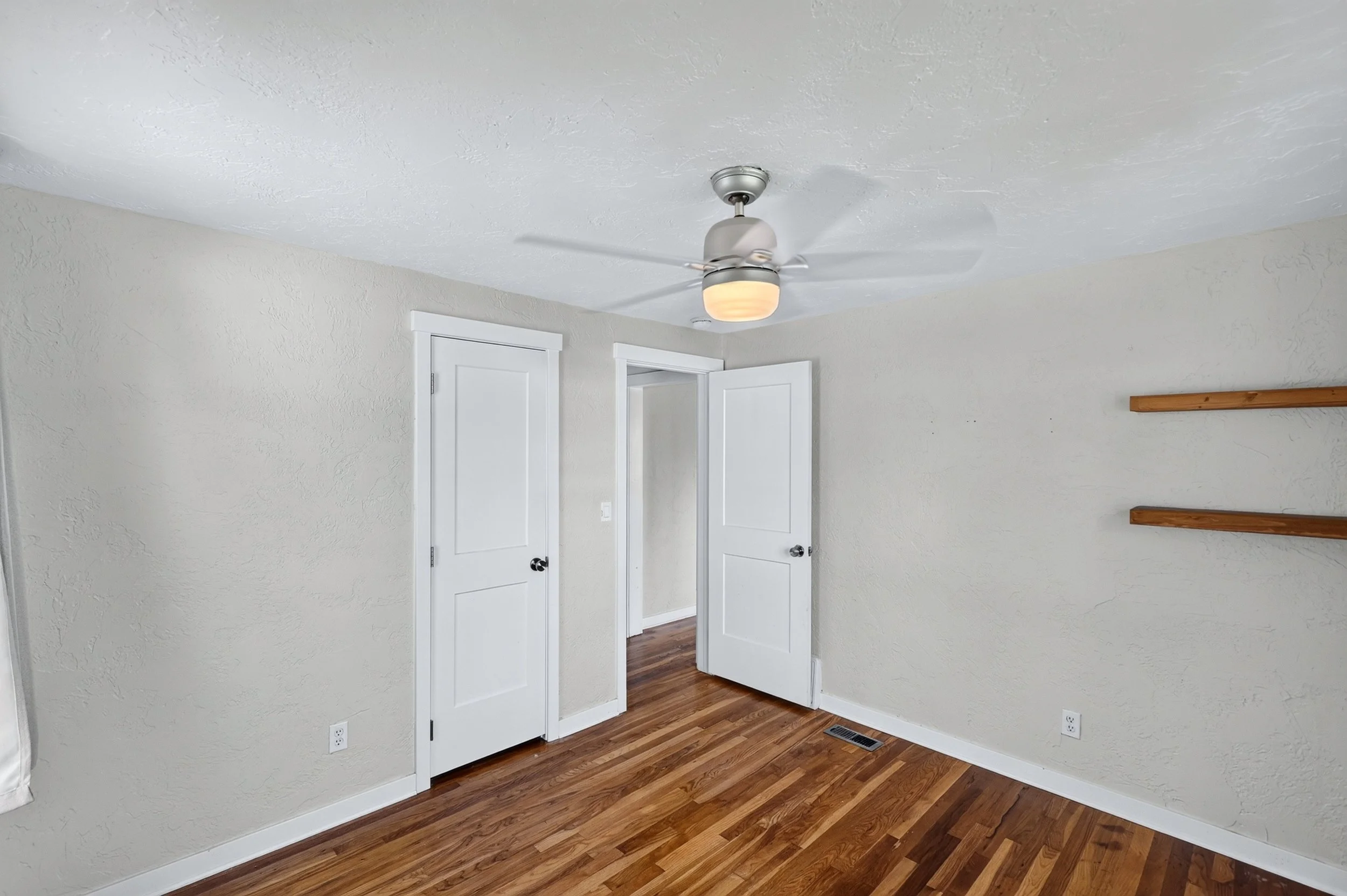 A room with light-colored textured walls, a wooden floor, a ceiling fan with light, a narrow doorway, and a wall-mounted wooden shelf.