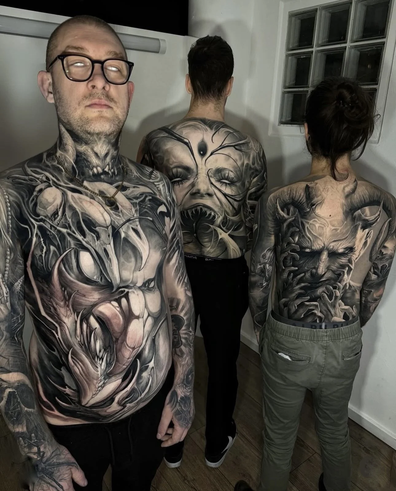 Three people with elaborate black and gray tattoos covering their upper bodies, facing away from the camera and showing detailed designs of faces, monsters, and abstract patterns.