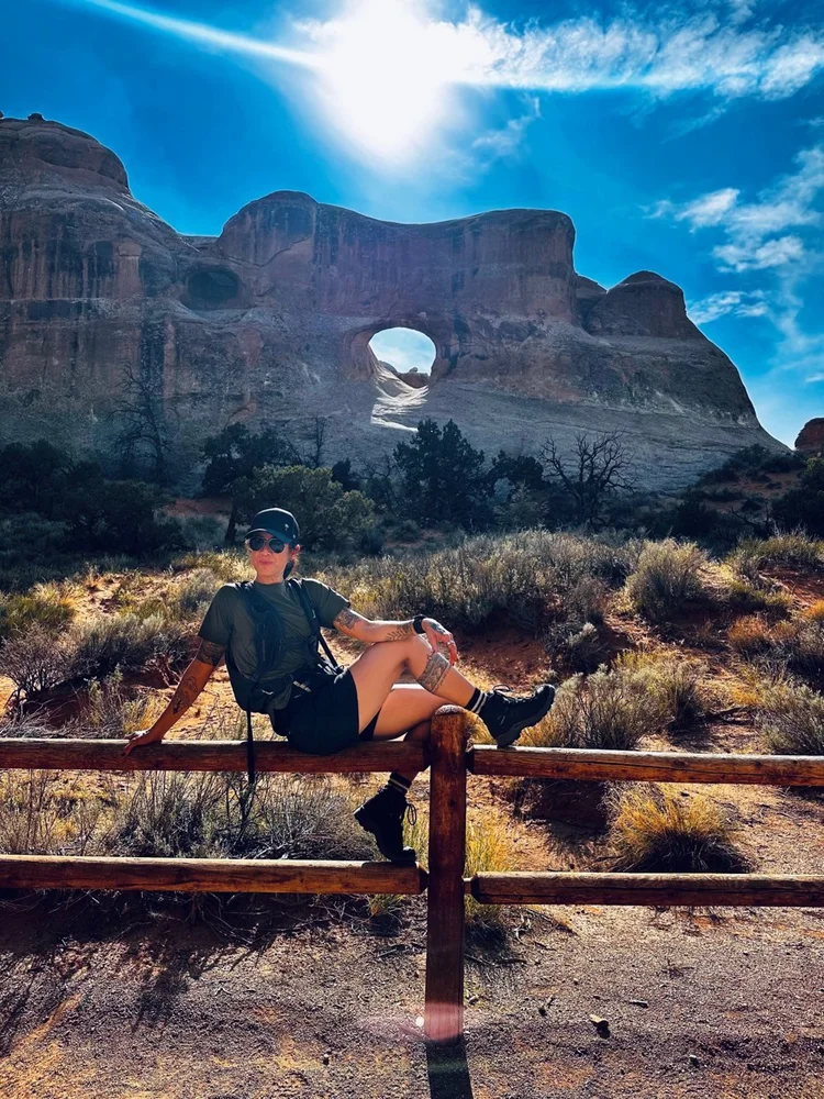 Cynthia Fowler sitting on a trail fence beneath a sandstone arch