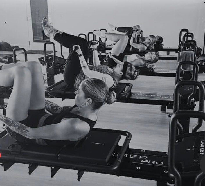Four women exercising on reformer Pilates machines in a gym, lying on their backs, lifting their legs and upper bodies.