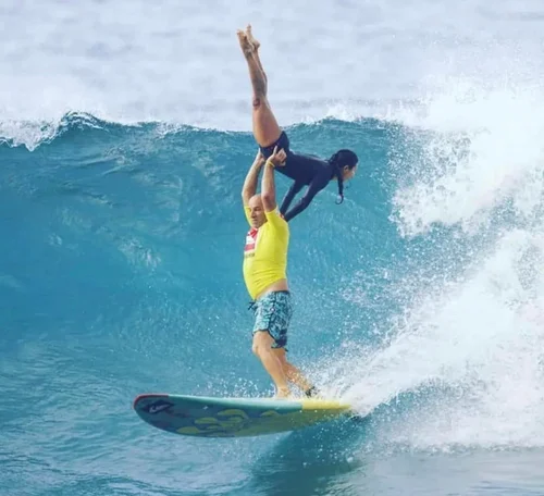Stephanie in a handstand on her partner's hands while tandem surfing a big wave at Mākaha