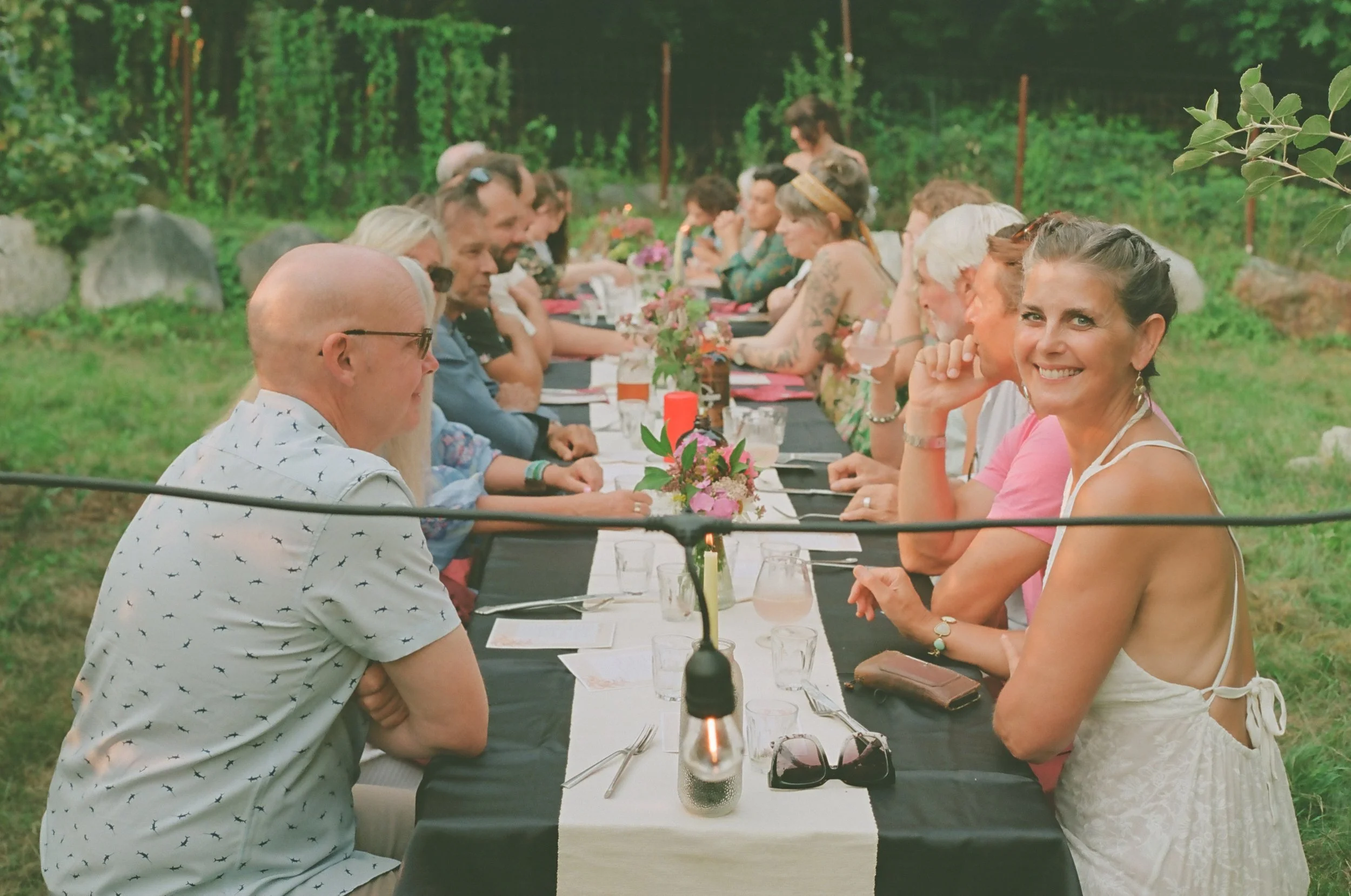 People seated at a long outdoor dinner table with a black tablecloth, surrounded by greenery, with floral centerpieces and candles.