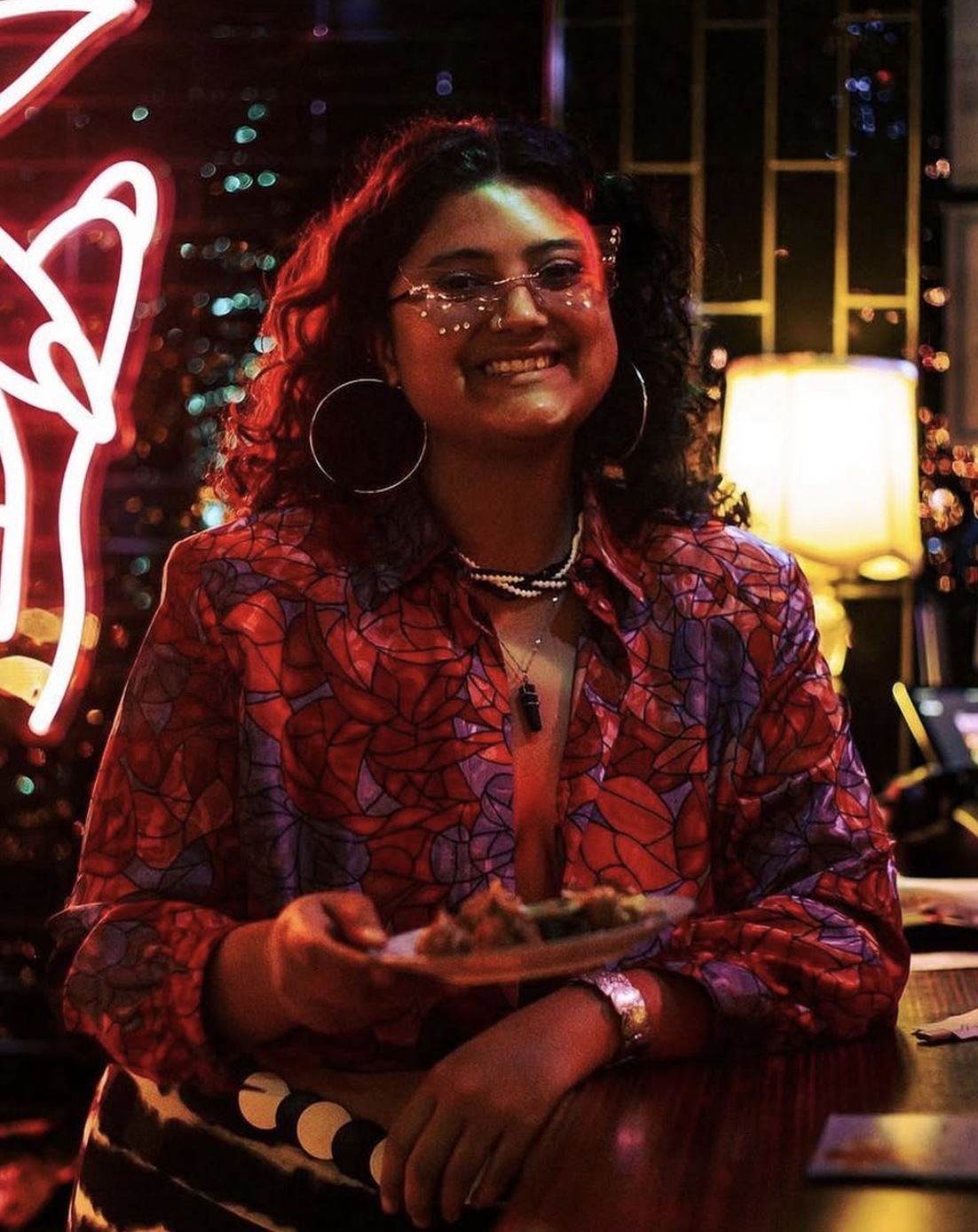 A woman, Asiah Pfaff, with curly hair and glasses smiles while holding a plate of food in a dimly lit, colorful restaurant with neon signs and warm lighting.