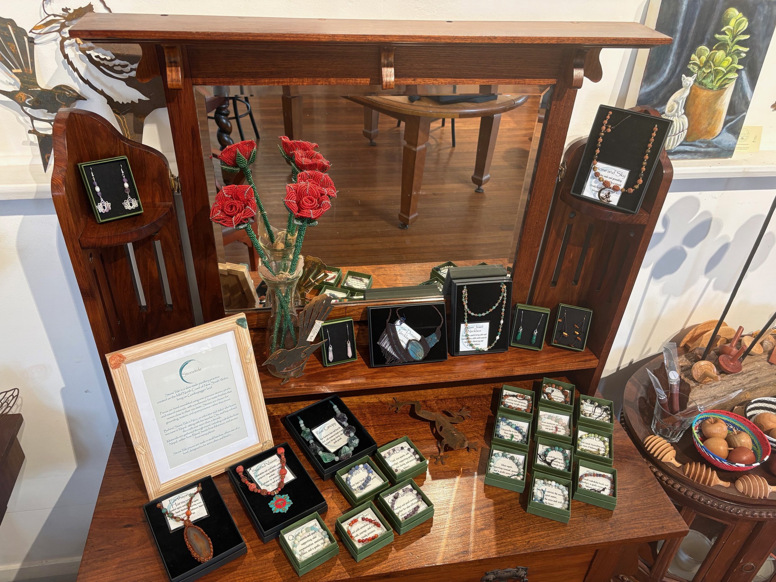 Display of handmade jewelry such as earrings, necklaces, and bracelets on a wooden table with a mirror, framed notes, and decorative items, including a glass vase with red roses and a wool craft flower arrangement.