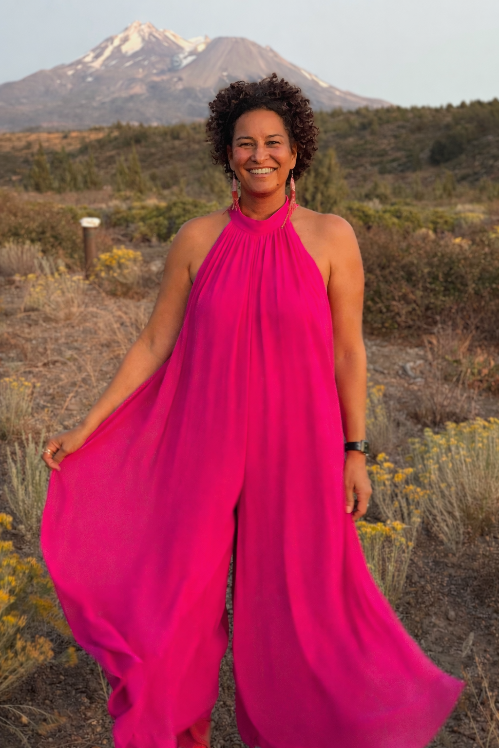 A woman in a bright pink dress standing outdoors in a desert landscape with yellow wildflowers, a mountain in the background, and a snow-capped volcano in the distance.