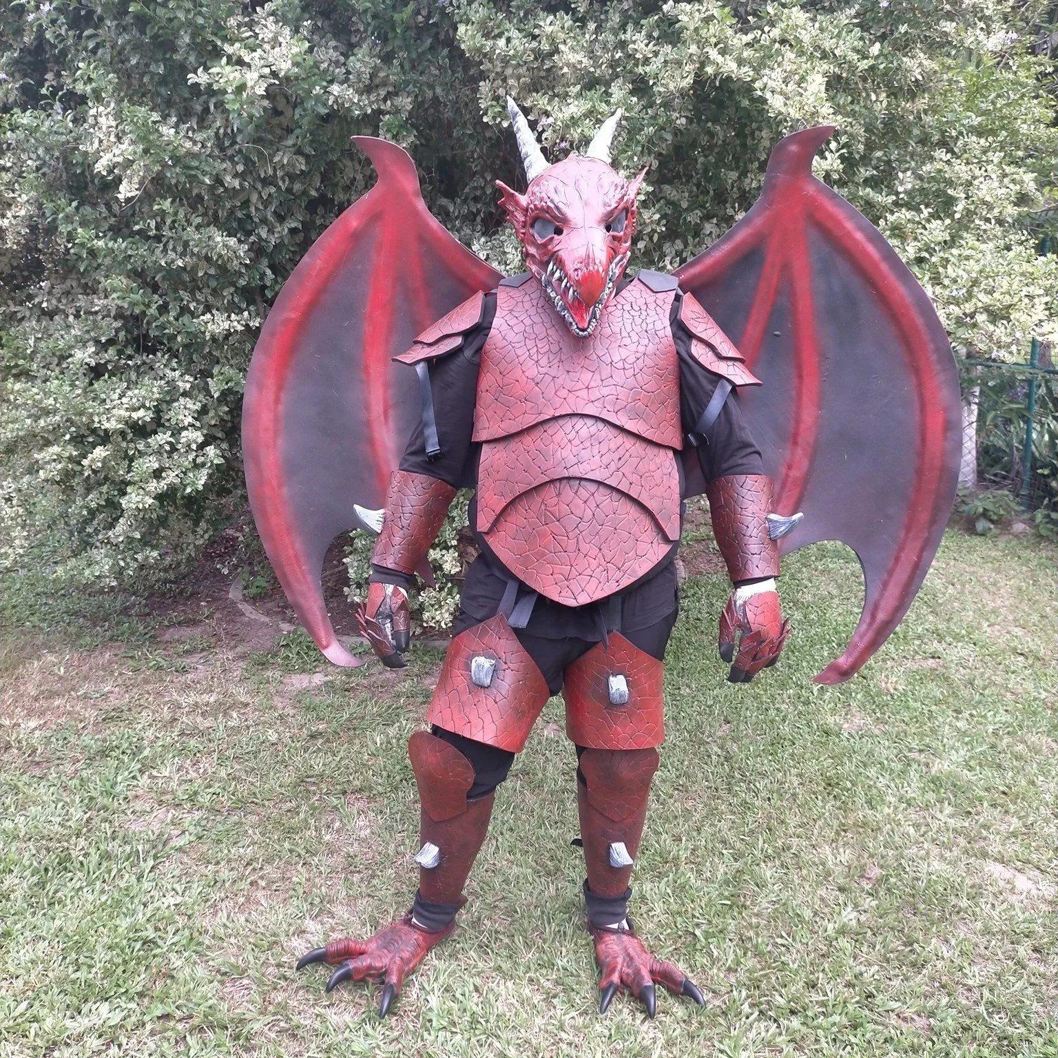 Person dressed in a red dragon costume with wings, claws, horns, and a detailed lizard-like face, standing outdoors on grass with bushes in the background.