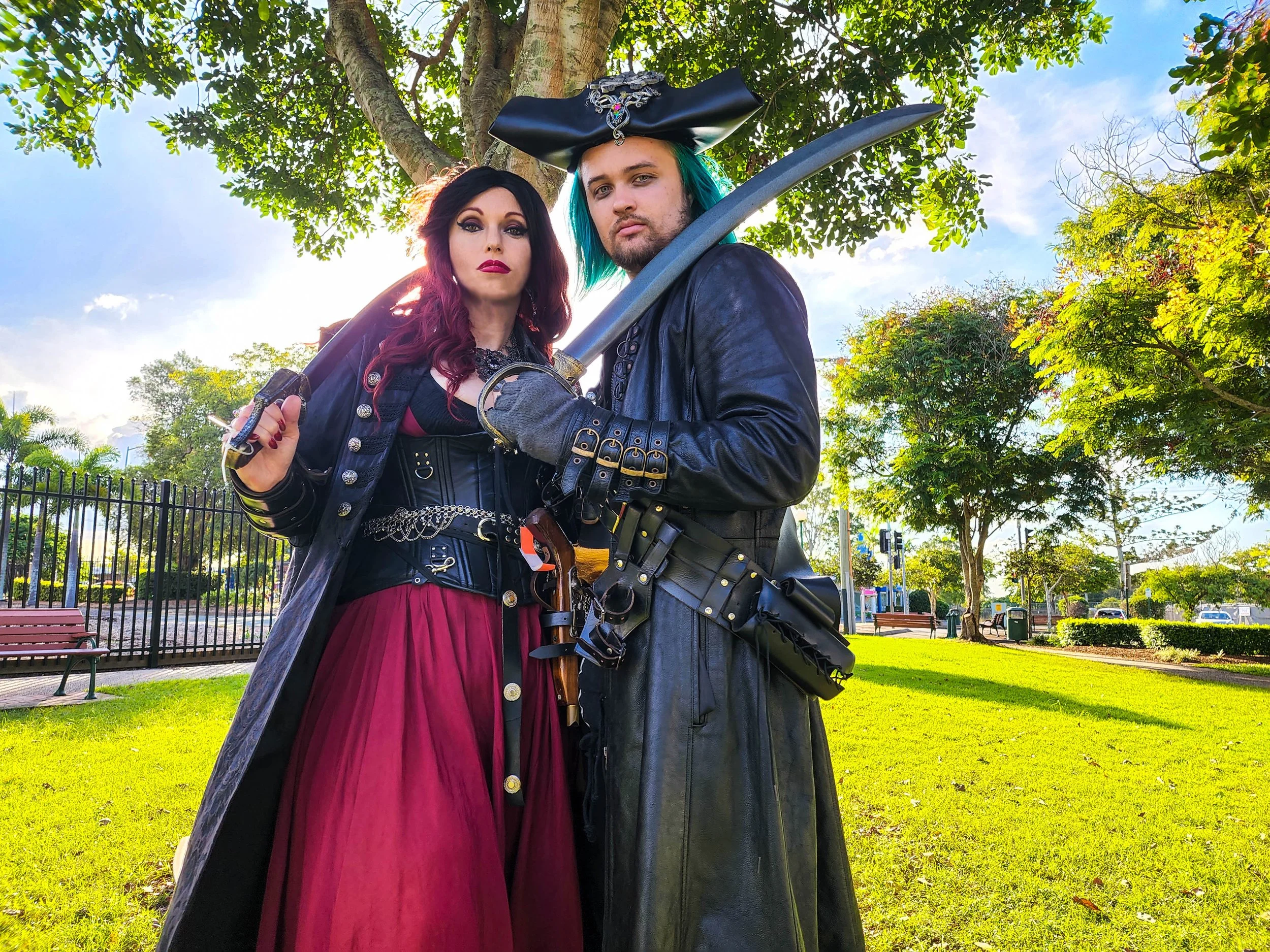 Two individuals dressed as pirates standing in a park with green grass, trees, and a clear sky in the background.
