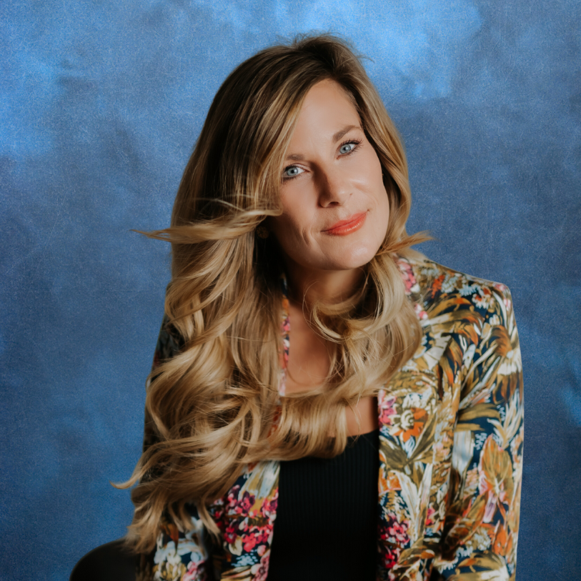 A woman with long, wavy blonde hair and blue eyes smiling slightly, wearing a colorful floral blazer and a black top, against a textured blue background.