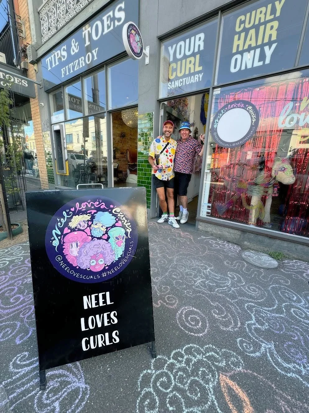Two men stand at the entrance of a hair salon called Neel Loves Curls, surrounded by colorful signs and chalk drawings on the sidewalk. One man wears a colorful polka dot shirt and shorts, and the other wears a patterned shirt and shorts.