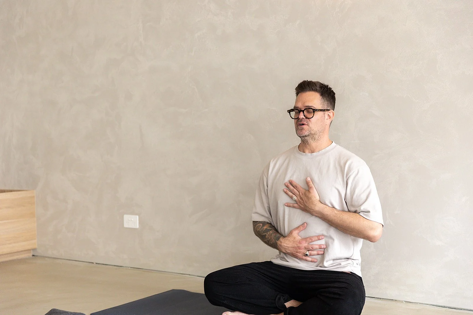 A woman and a man practicing meditation in a bright room with white brick walls, sitting cross-legged with eyes closed and noses pinched shut.