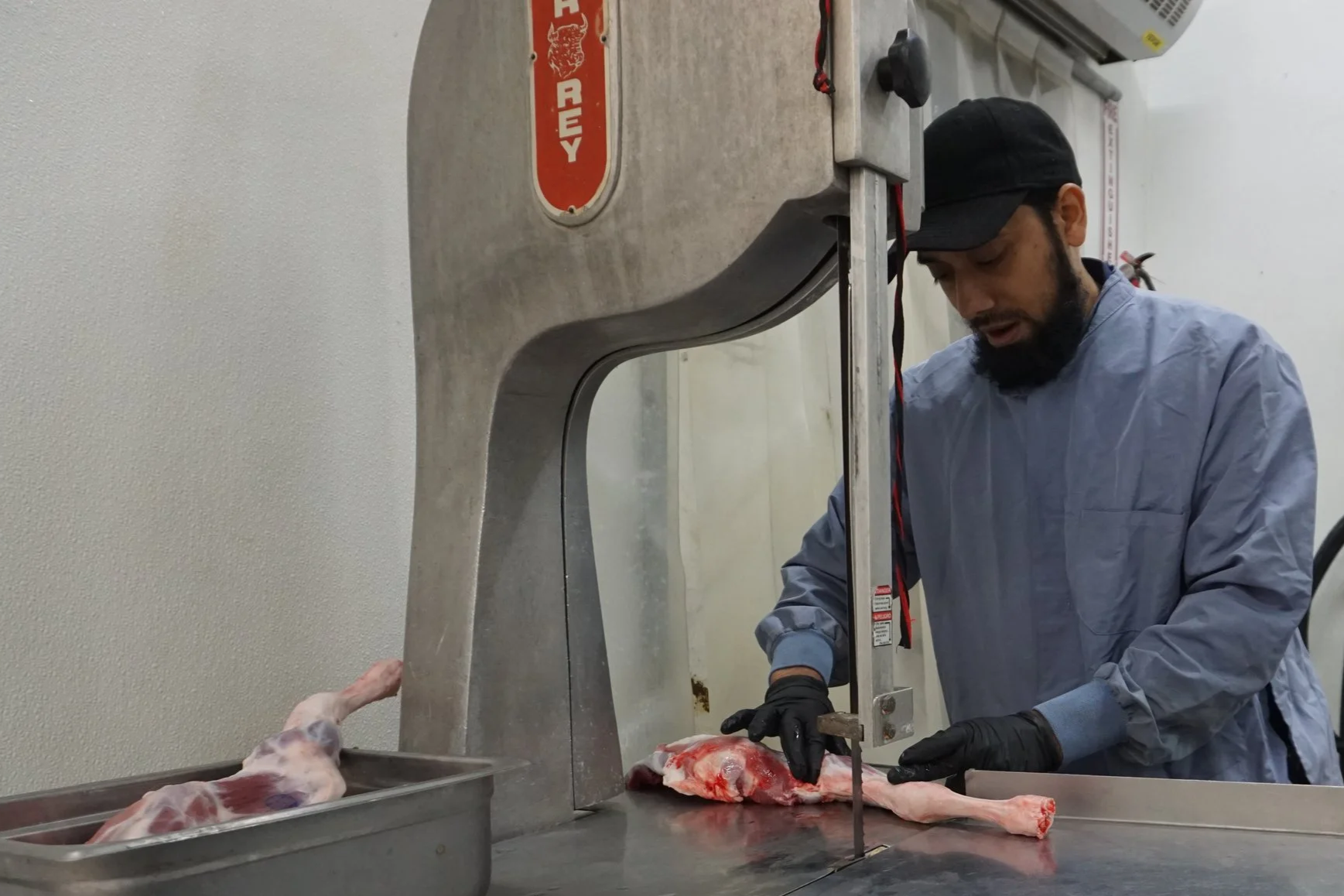 A man dressed in a gray coat, black gloves, and a black cap is working with a saw on a large piece of meat on a stainless steel table, with a meat saw and other equipment visible in the background.
