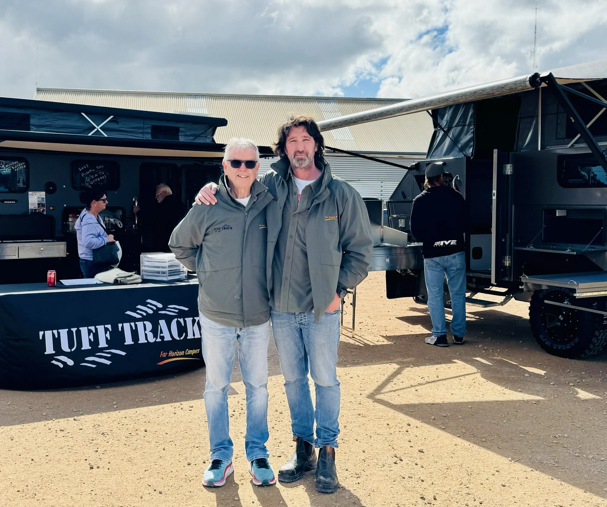 Two men standing outside, smiling, with a black table that says 'Tuff Track' behind them, and a large truck or RV with a canopy. The background shows a cloudy sky and a large building.