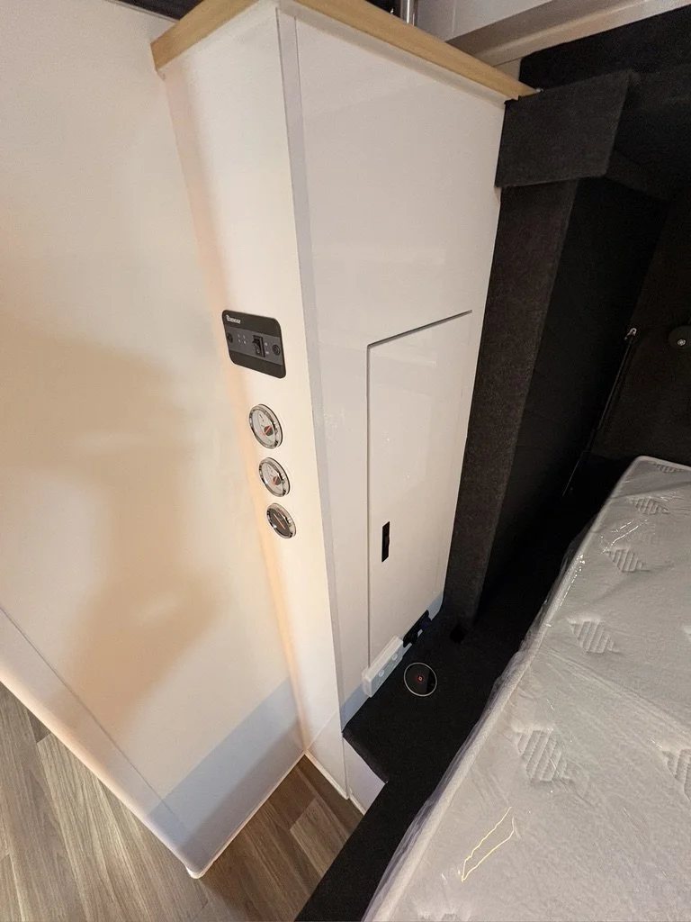 View of a modern bedroom corner with a white wardrobe, black bedside table, and a bed with a mattress, along with some control panels and gauges on the wardrobe.