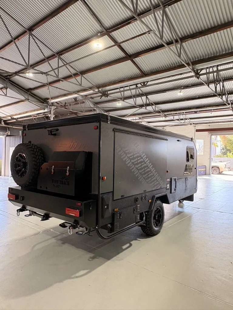 A black, off-road camper trailer with a spare tire mounted on the back inside a large indoor warehouse with a high ceiling and metal beams.