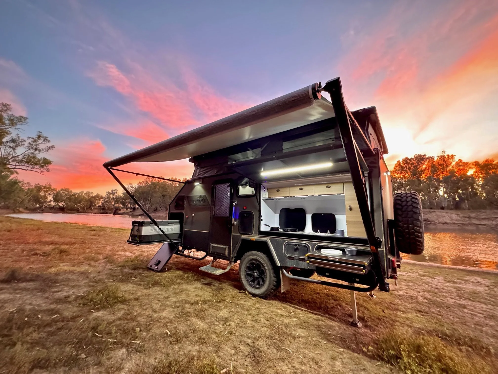 A black off-road camper trailer with an extended awning set up along a riverbank at sunset, surrounded by trees with a colorful sky.