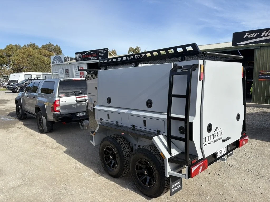 A gray toy hauler trailer attached to a gray Toyota Land Cruiser in a parking lot with other vehicles and buildings in the background.
