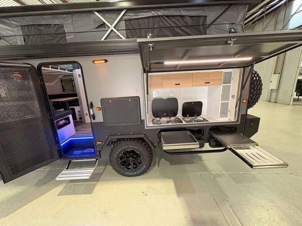 A compact outdoor camper trailer with kitchen and storage amenities, including a slide-out stove, sink, and refrigerator, located inside a warehouse.