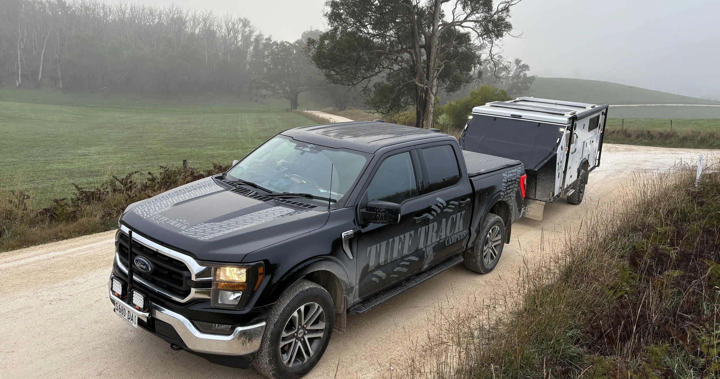 Black Ford truck towing a camper on a dirt road in a foggy rural landscape with grassy fields and trees.