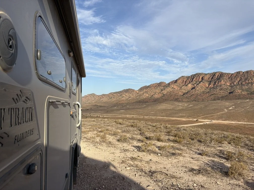 Side of a travel trailer in a desert landscape with mountains in the distance and a partly cloudy sky.