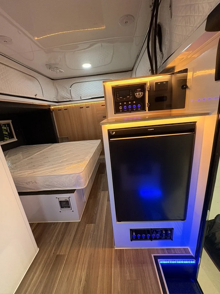 A small, modern interior of a camper or tiny house with a bed, microwave, mini refrigerator, and control panels, illuminated by warm LED lighting.