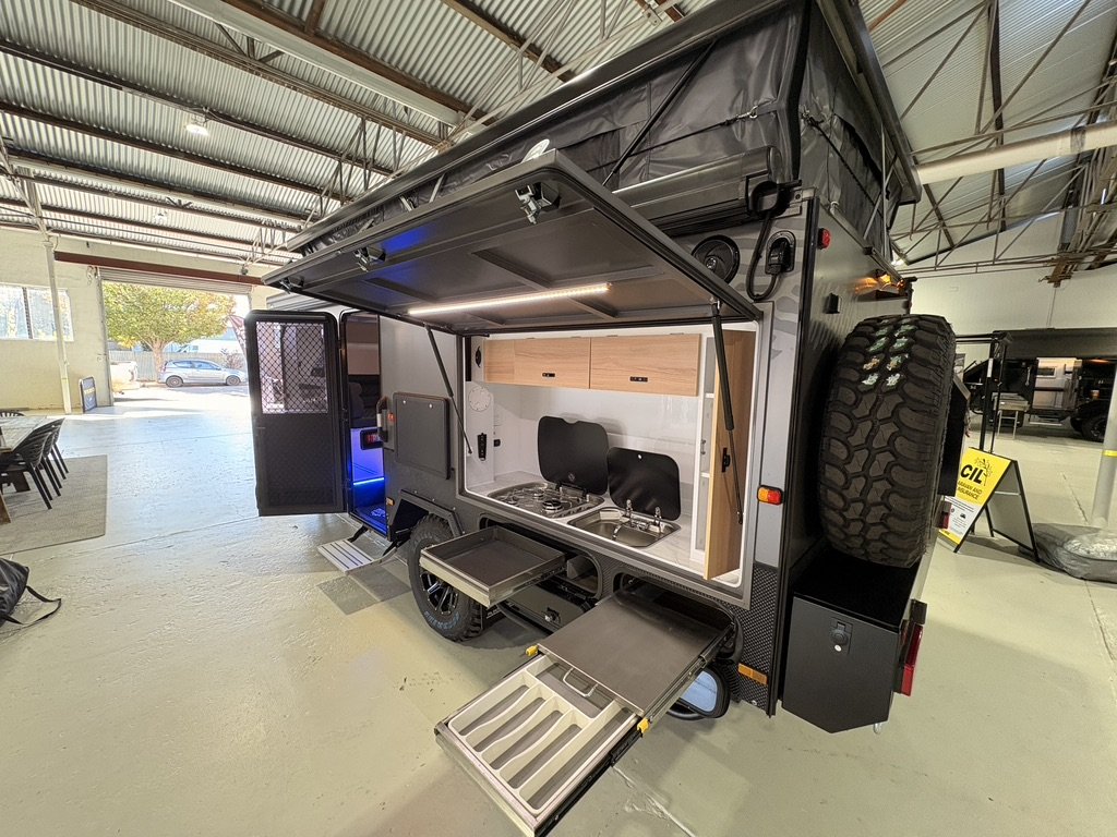 A compact camper trailer with a slide-out kitchen area, including a stove, sink, and cabinets. The trailer is inside a spacious warehouse with other trailers in the background.