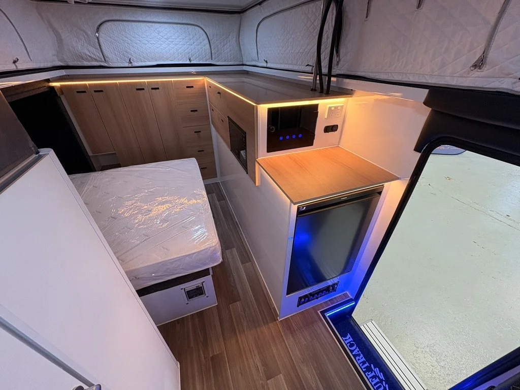 Interior of a compact camper van with a bed, wooden cabinets, a small refrigerator, and a countertop with LED lighting.