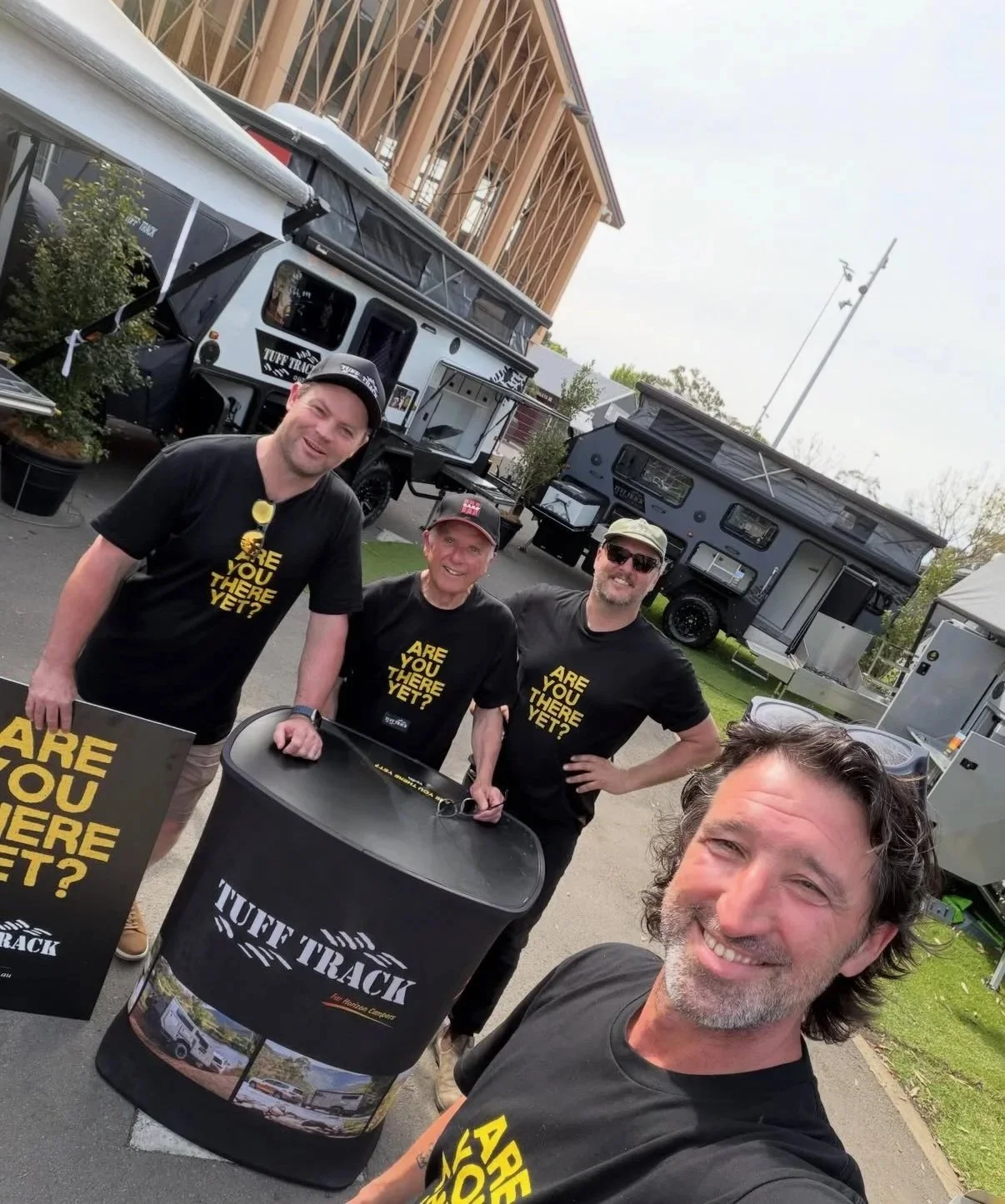 Four men posing outdoors in front of a display of two large off-road travel trailers, with banners that read 'Are You There Yet?' and 'Tuff Track'. The man in the foreground is taking a selfie. All are wearing black T-shirts with yellow text and appear happy.