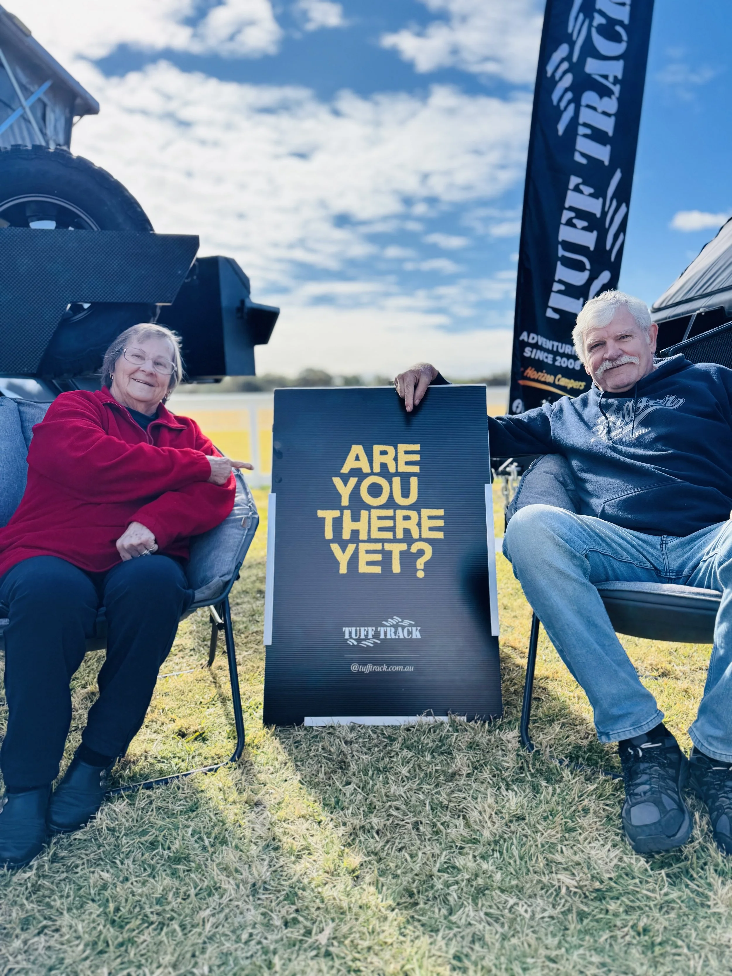 An elderly woman and man sitting outdoors on foldable chairs next to a sign that reads 'Are You There Yet?'. The background shows a partly cloudy sky and a large black flag with white text.
