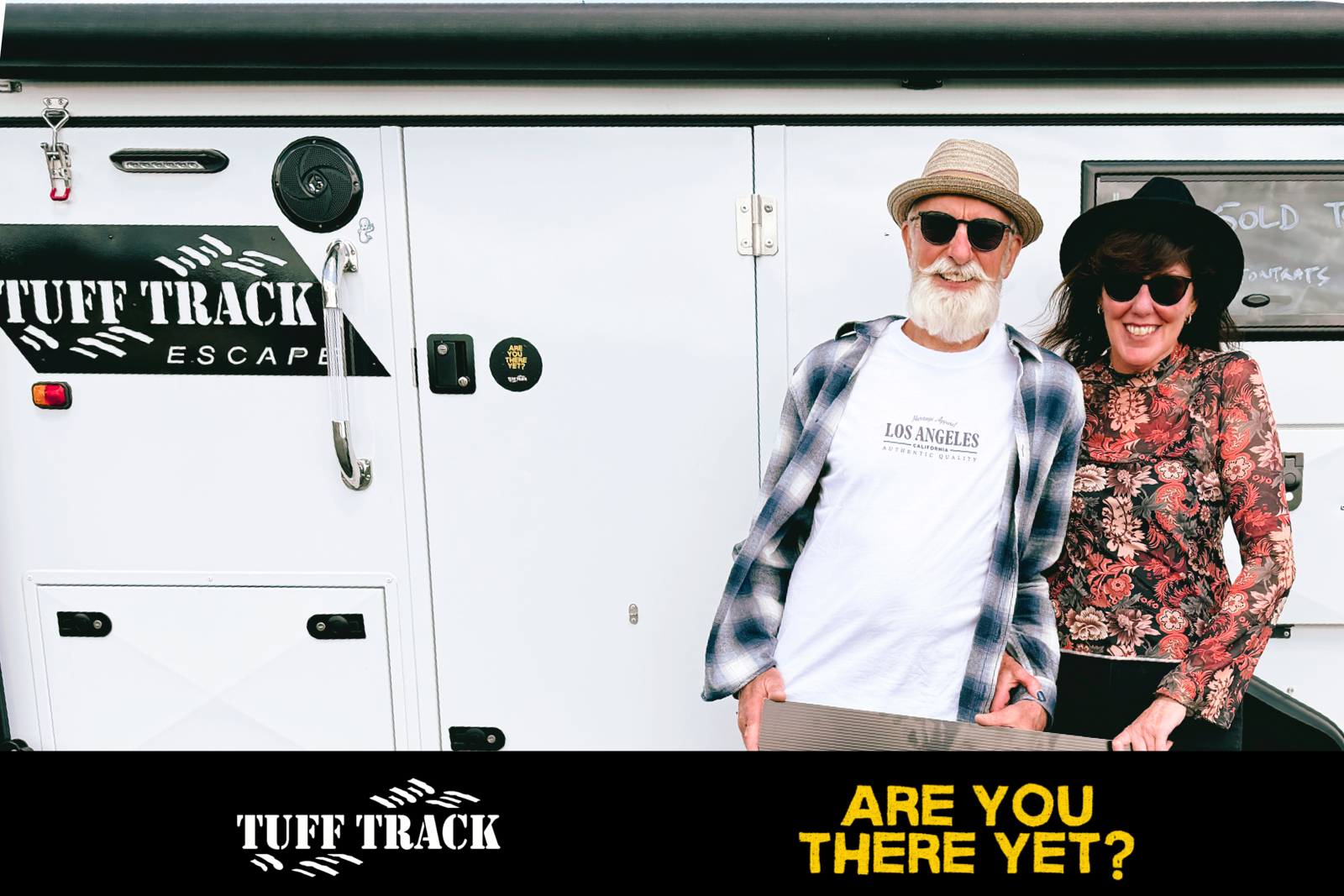 Two smiling people, a man with a white beard in sunglasses, a hat, and a casual shirt, and a woman in sunglasses and a floral top, standing in front of a white trailer with the logo 'TUFF TRACK ESCAPE' and the text 'Are you there yet?' at the bottom.
