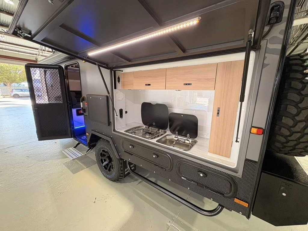 Interior of a compact black off-road camper with a kitchen area, including a sink, stove, and wooden cabinets, located inside a warehouse.