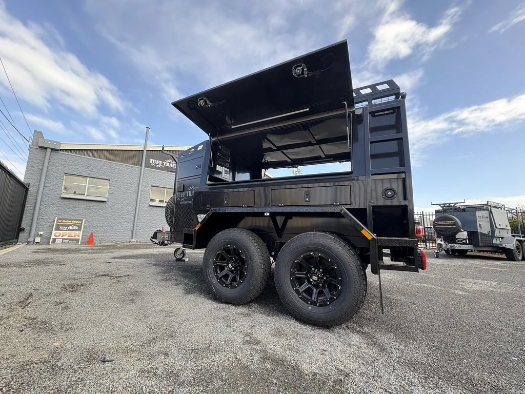 Black off-road trailer with storage and a large open hatch, parked in a lot with a gray building and other trailers in the background under a partly cloudy sky.
