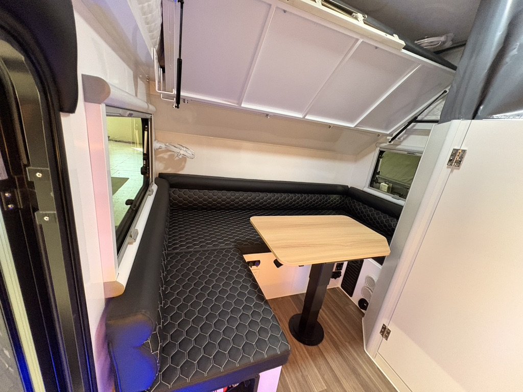 Interior of a compact camper with black cushioned seating, a small wooden table, and windows.