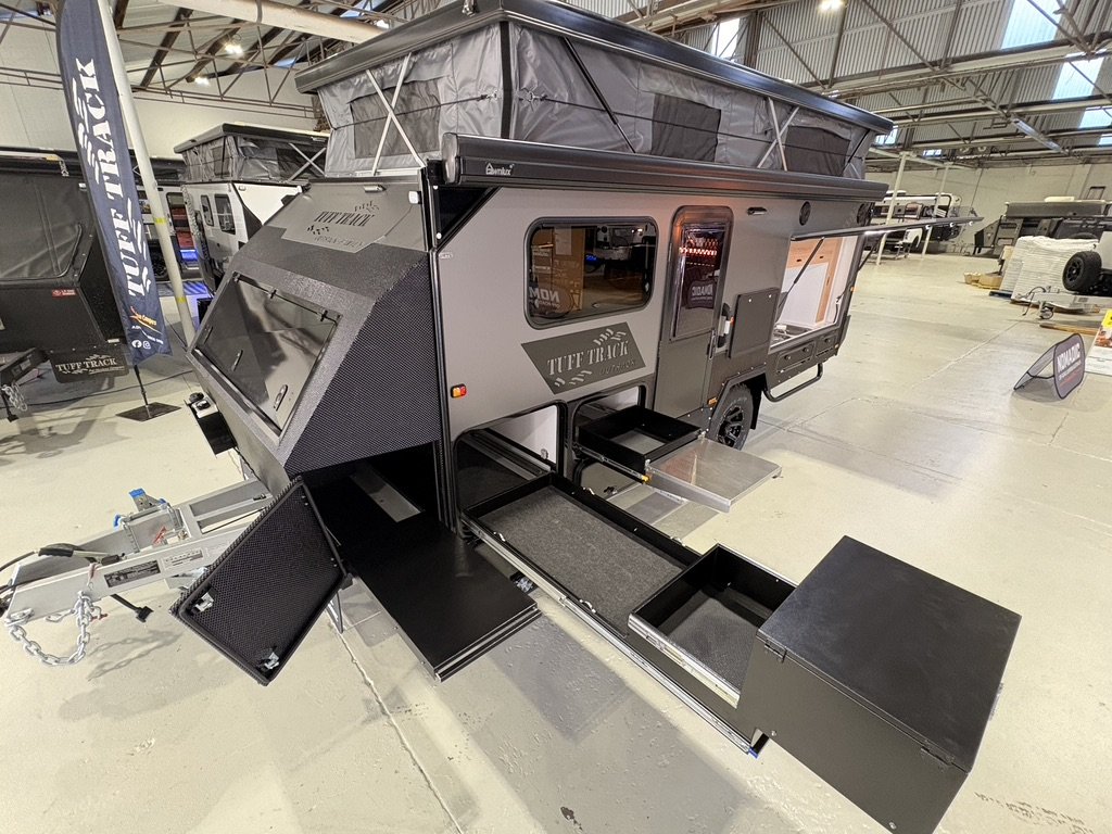 A collapsible travel trailer with slide-out extensions and outdoor storage compartments displayed at an indoor exhibition.