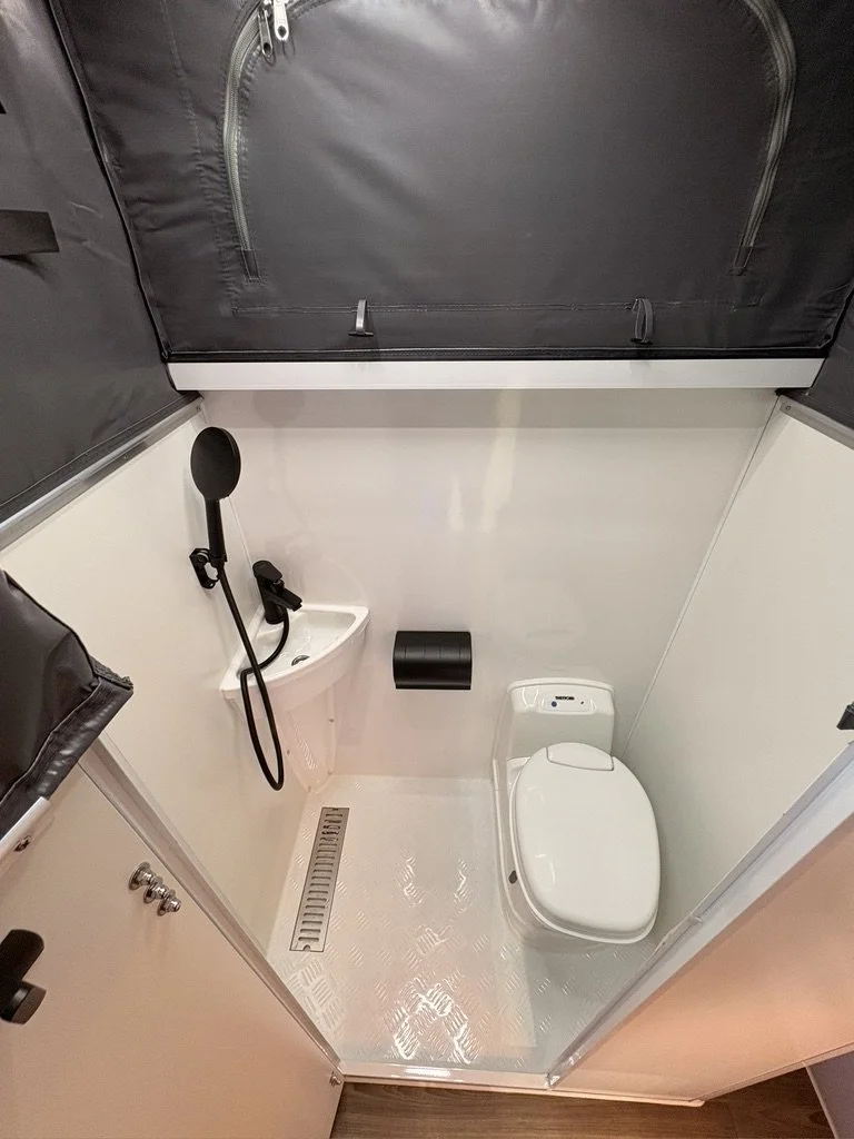 Small bathroom with a toilet, a small sink with a showerhead attached, a black toilet paper holder, a metallic floor drain, and a gray cabinet with a zippered door.