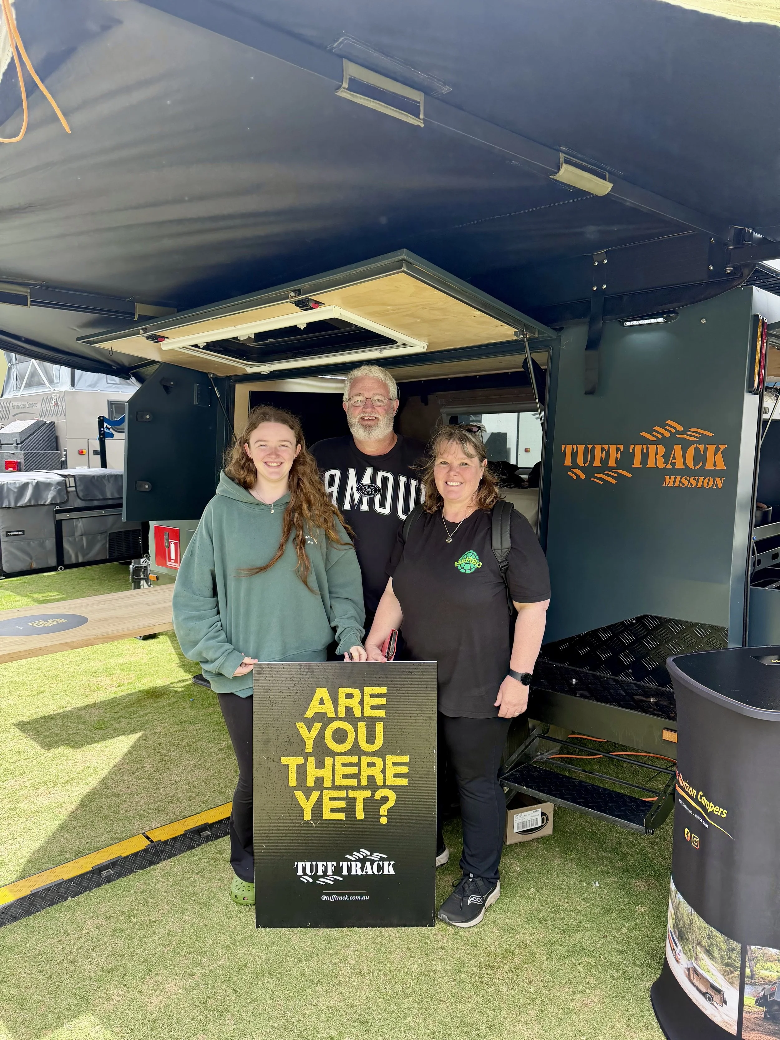 Three people standing behind a sign that says, "ARE YOU THERE YET?" at an outdoor event, with a black trailer labeled "TUFF TRACK MISSION" and a black display stand to the right.