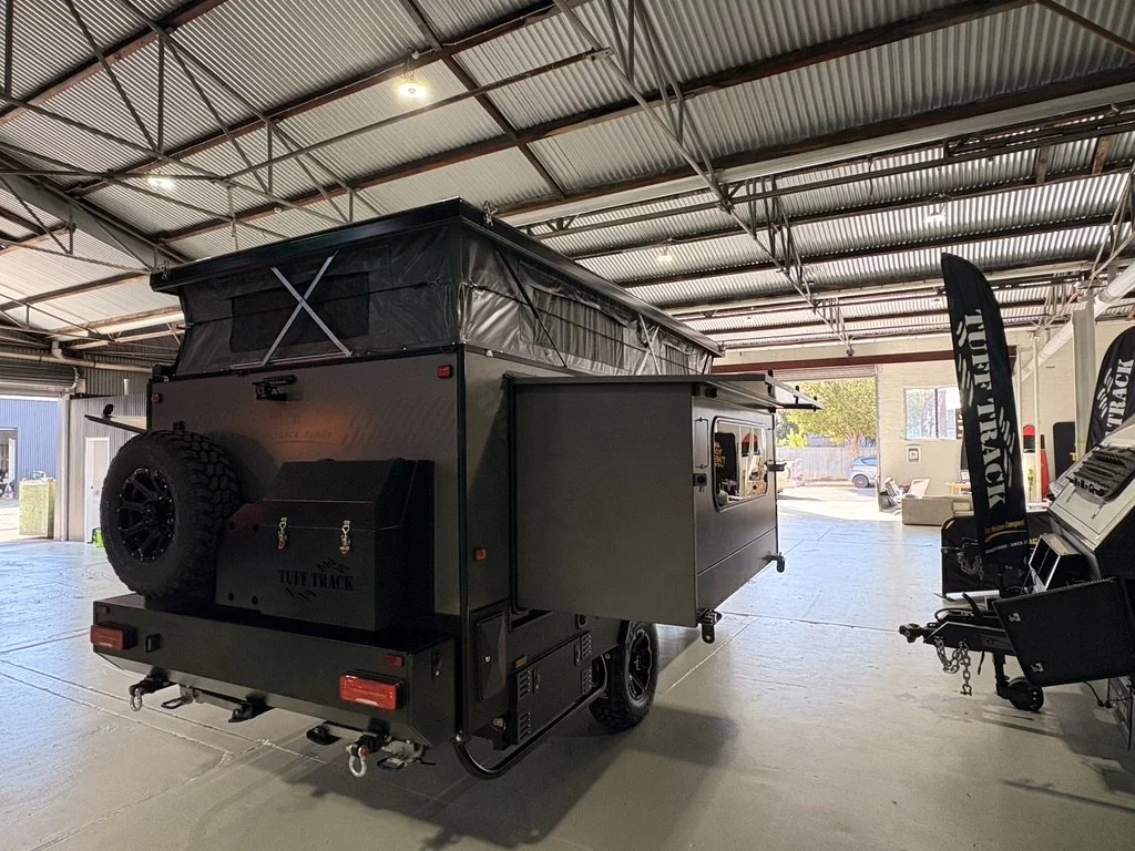 A black off-road camper trailer with a spare tire mounted on the back, interior lighting, and fold-out side panels, parked inside a spacious warehouse with a metal roof and large open doors.