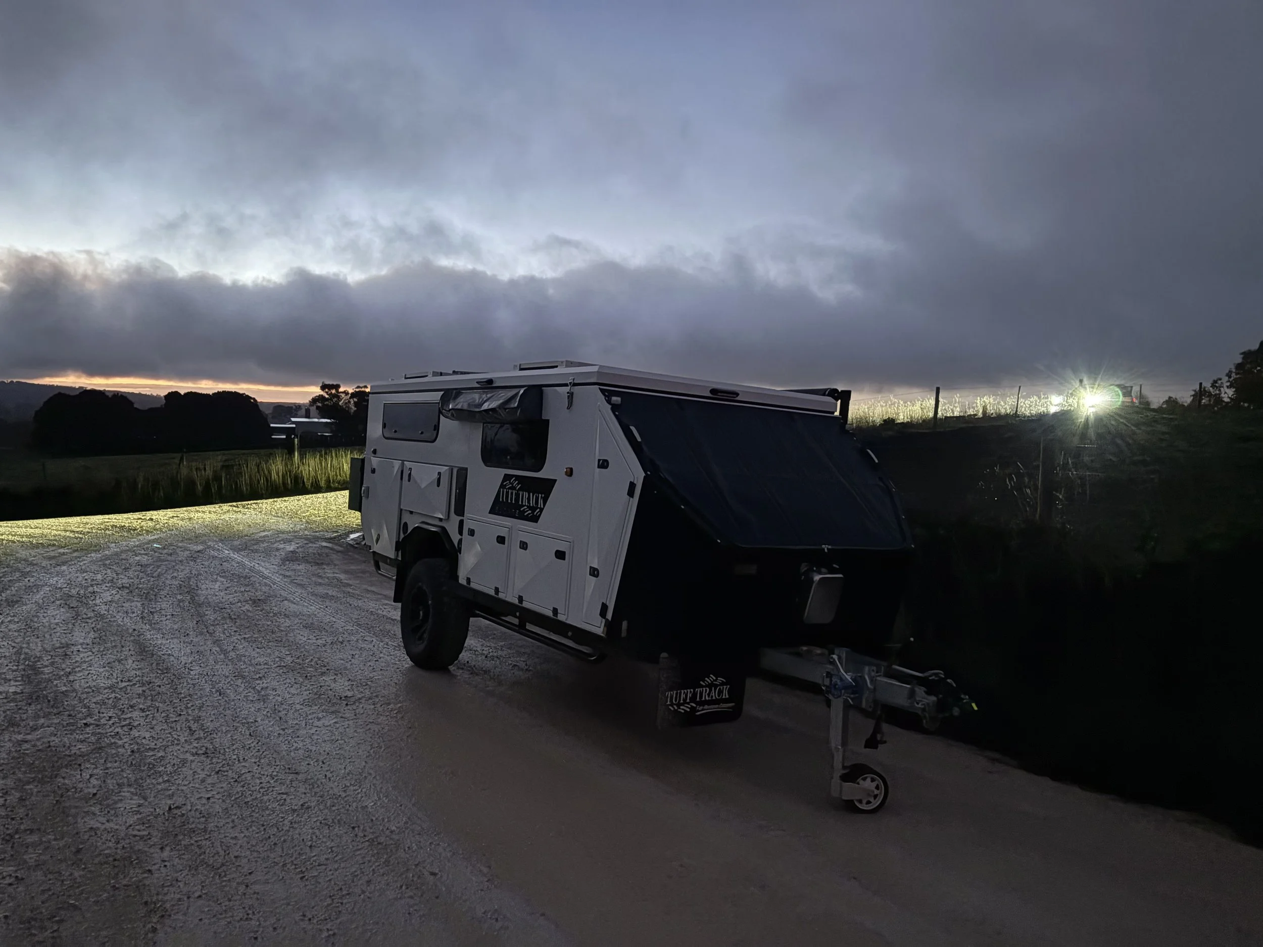 White off-road camper trailer parked on a gravel road at dusk with dark clouds overhead and a green glow from the horizon and distant headlights.