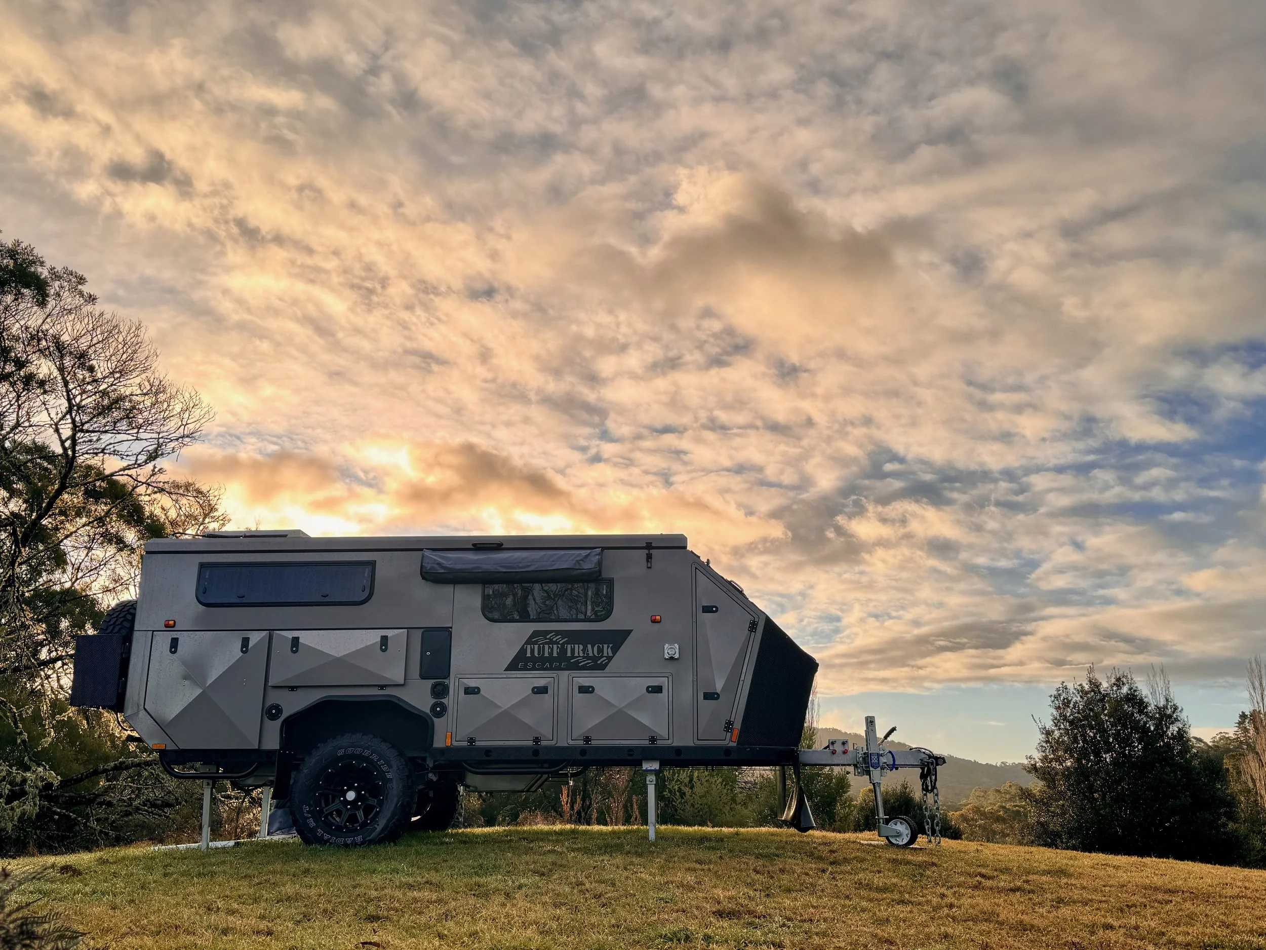 A gray and black off-road camper trailer parked on a grassy hill under a partly cloudy sky at sunset.