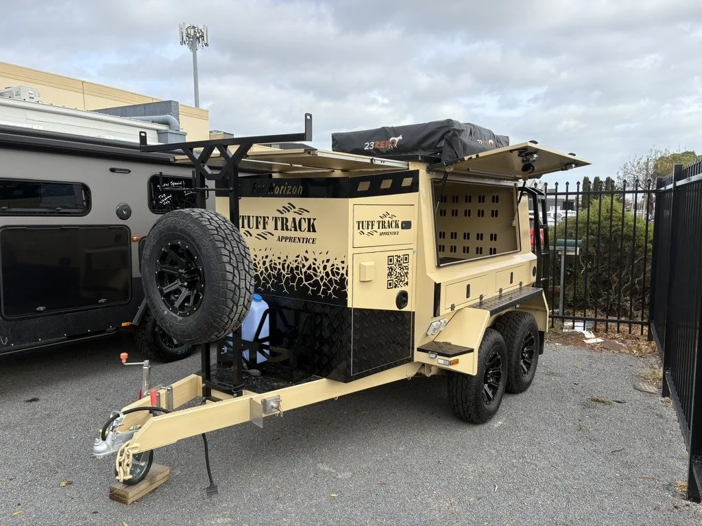 A beige off-road trailer with black accents parked on a paved lot, equipped with a spare tire on the front, an adjustable awning, storage compartments, and a roof-mounted storage box.