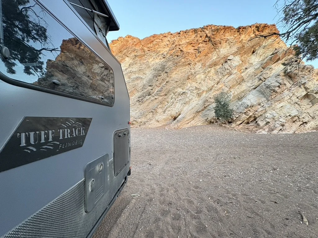 A photo taken from near a Flinders 16ft hybrid camper in South Australia "TUFF TRACK FLINDERS," parked on a dirt ground next to a rocky cliff with sparse vegetation, under a clear blue sky.