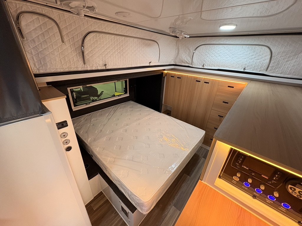 Interior of a compact camper or RV with a bed, wooden cabinetry, and control panel, illuminated with warm LED lighting.