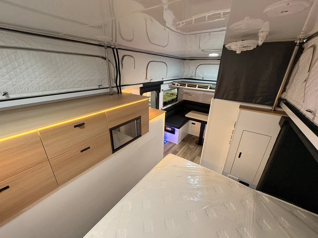 Interior of a compact living space, possibly a tiny home or camper, with wooden cabinetry, a bench with table, small kitchen area, and sleeping area with a fold-out bed or bunk bed.