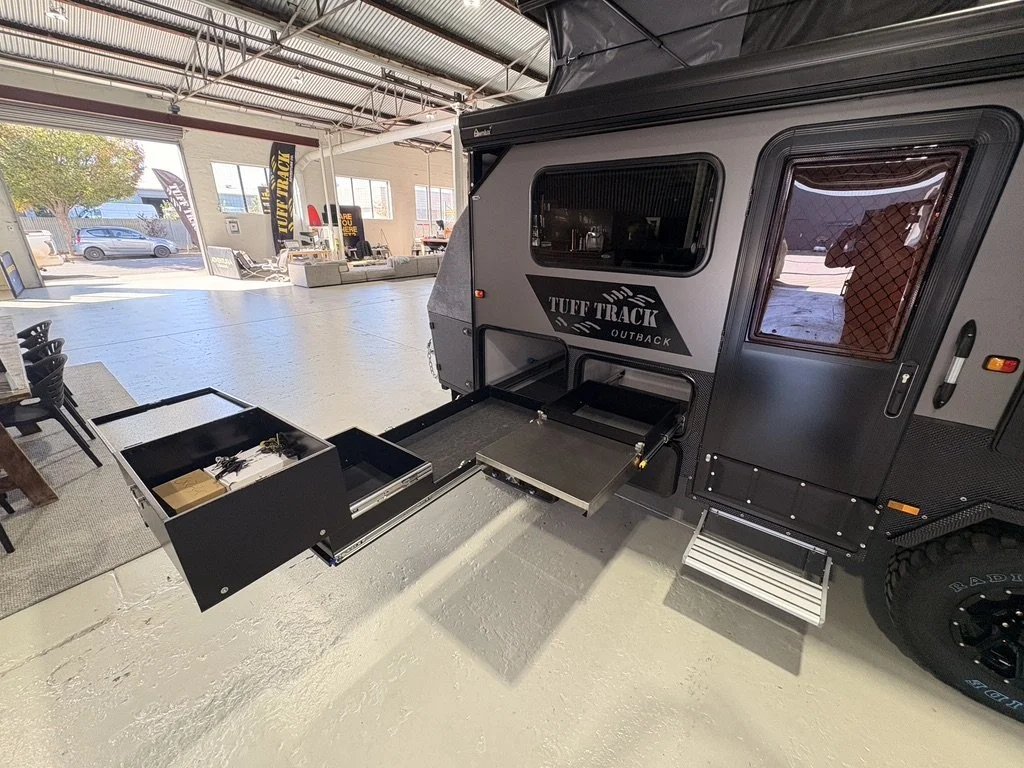 An off-road travel trailer named TUFF TRACK OUTBACK inside a showroom, with an open pull-out storage drawer and a fold-out step.